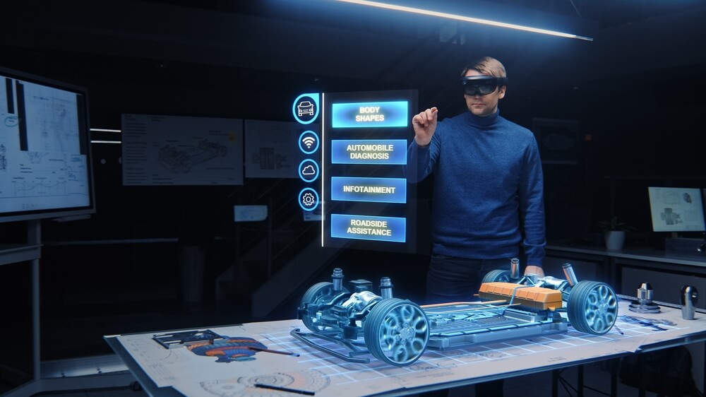 A man in a blue sweater stands before a computer screen displaying a car model