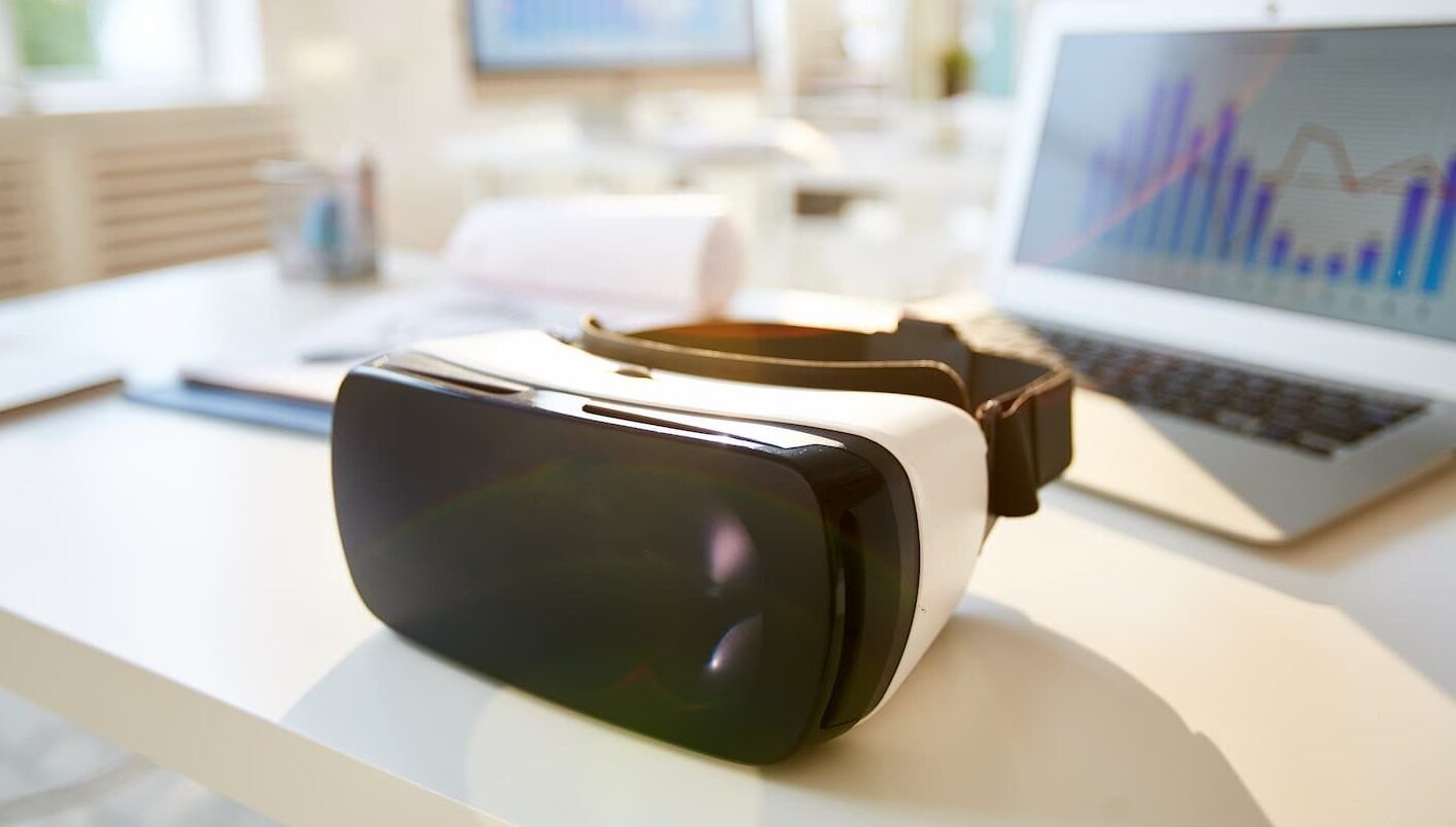 A VR headset on a modern desk, next to a laptop displaying financial charts and data.